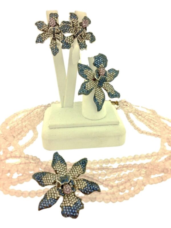 A set of Orchid Brooch - Sapphire jewelry including earrings and a necklace with light pink beads and blue flower pendants, displayed on a white stand.