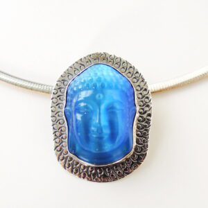 A silver necklace with a large, oval-shaped Buddha Silhouette brooch / pendant featuring a blue, embossed face against a textured silver background.