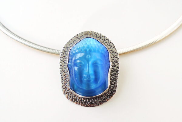 A silver necklace with a large, oval-shaped Buddha Silhouette brooch / pendant featuring a blue, embossed face against a textured silver background.