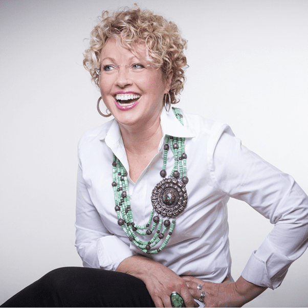 Smiling woman with curly blonde hair, wearing a white shirt and large green beaded necklace, sitting against a light gray background.